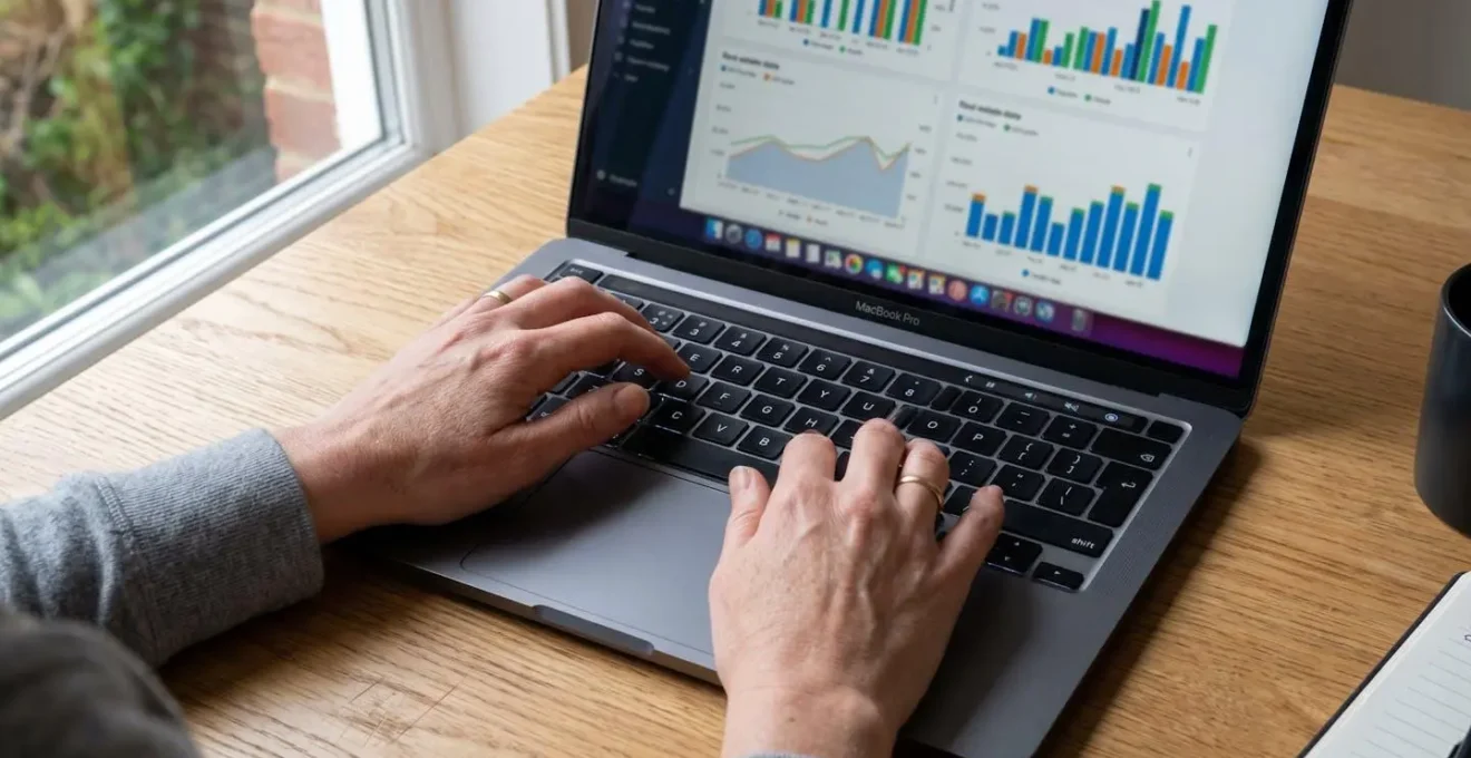 Gros plan sur des mains tapant sur un clavier d'ordinateur portable moderne, avec un reflet d'écran montrant un graphique de données floues sur un bureau en bois clair